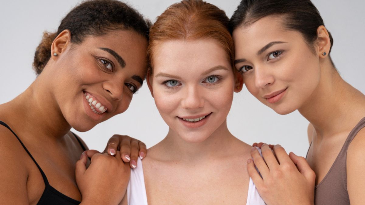 Três mulheres de tons de pele distintos com cabelo preso e sorrindo